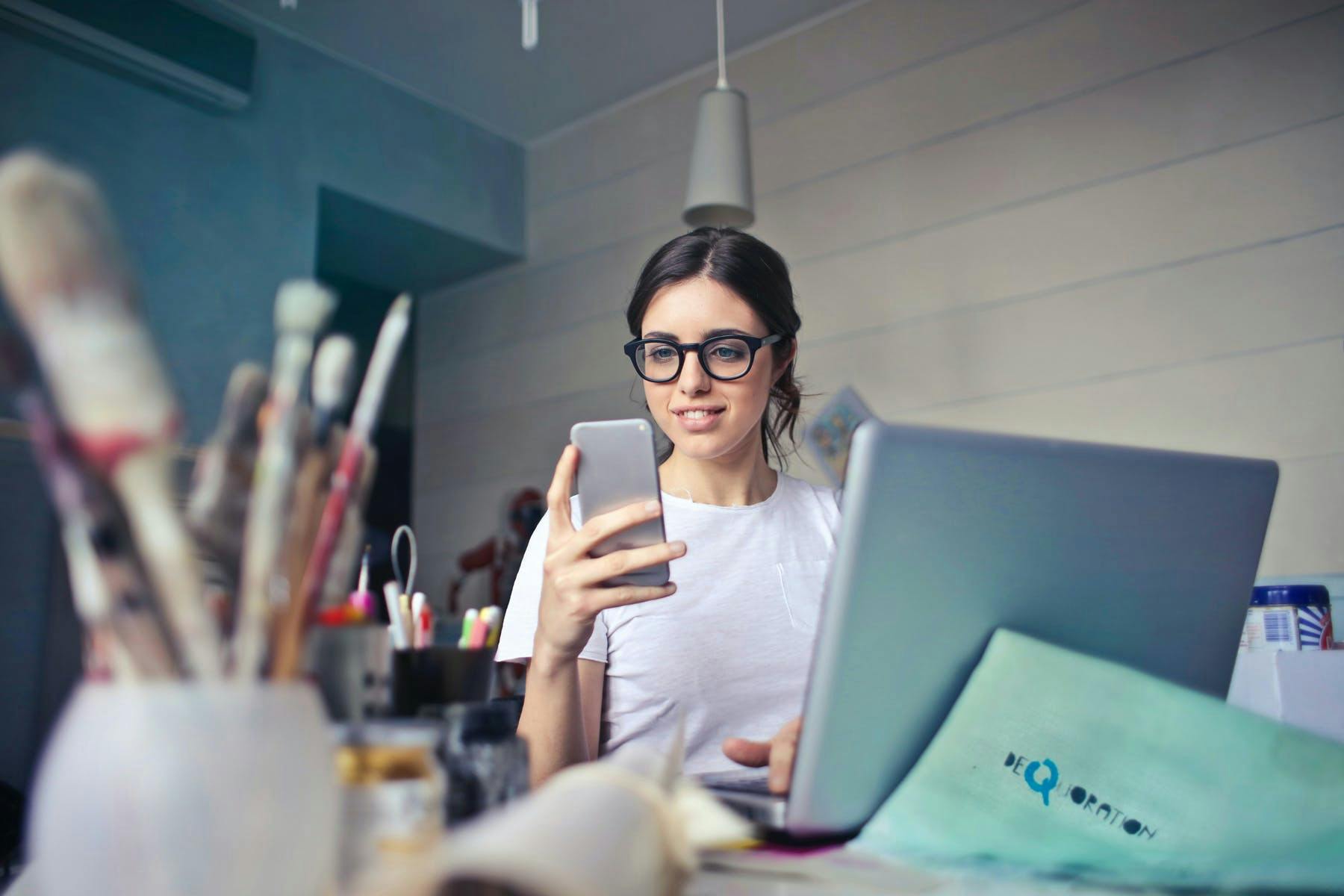 A young freelance designer with glasses looking at her iPhone at a desk with a laptop and paint brushes. 
