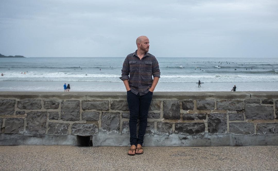Stefan Feser, a young web development teacher posing at a sea-side with surfers in the background. 